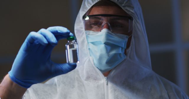 Scientist in full protective gear holding and examining a vial in a laboratory. Suitable for use in articles or advertisements related to medical research, biotechnology, healthcare settings, and scientific advancements. Can also be used for content focused on lab safety, pandemic response, and vaccine development.
