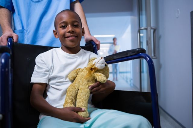 Portrait of boy sitting on wheelchair and holding teddy bear in corridor at hospital