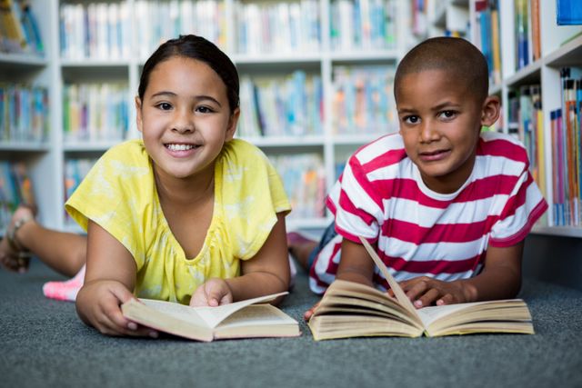 Two children lying on the floor in a library, reading books together. They are smiling and appear to be enjoying their books. The scene evinces a sense of learning, friendship, and happiness. Ideal for educational materials, library promotions, and children's books. Captures the essence of literacy, diversity, and childhood joy.
