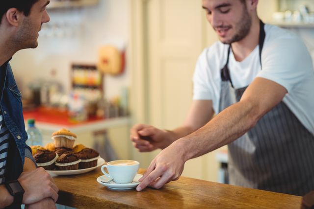 Barista serving a cup of coffee to a young customer at a cozy cafe. Ideal for use in articles or advertisements related to coffee shops, customer service, hospitality industry, and small businesses. Can also be used in promotional materials for cafes and bakeries.