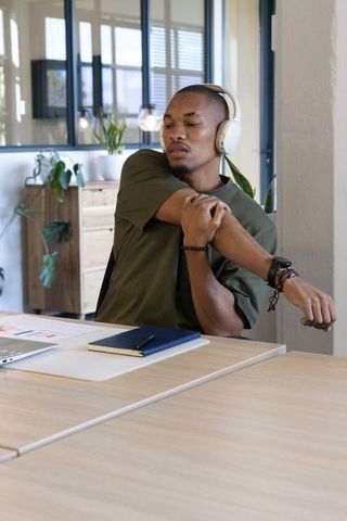 African american man stretching at desk wearing headphones while working in modern office