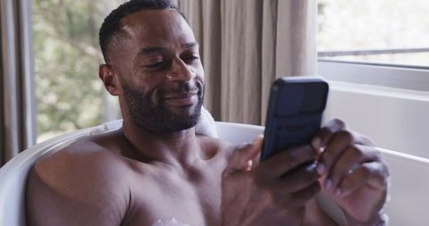 Middle-aged man relaxing in bathtub using smartphone near window natural light
