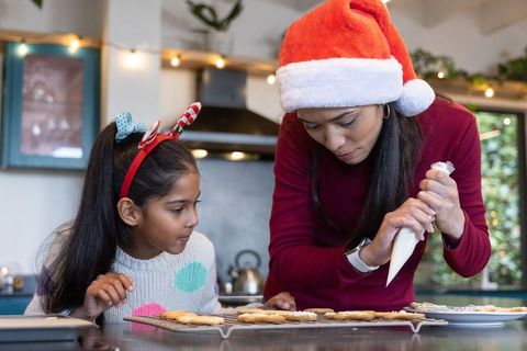 Indian mother wearing santa hat decorating christmas cookies with daughter in cozy kitchen