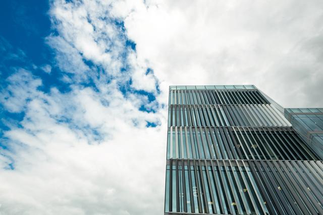 This image captures a modern office building with a glass facade from a low angle, set against a blue sky with scattered clouds. It is ideal for use in corporate presentations, real estate promotions, architectural portfolios, and business websites to convey professionalism and contemporary urban life.