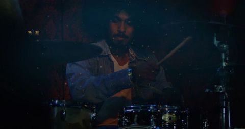Playing indian drummer striking drum kit and cymbals on moody dimly lit stage wearing denim jacket
