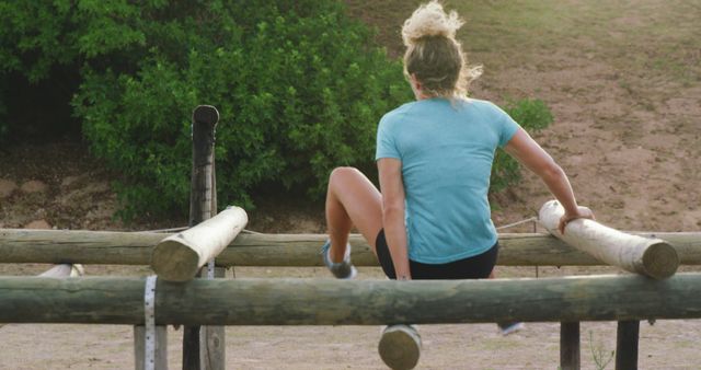 Back view of a woman in blue shirt and black shorts navigating an outdoor obstacle course with wooden logs, set in a natural environment. Ideal for themes of fitness, adventure, teamwork, or nature-inspired challenges. Perfect for promoting outdoor activities, athletic training programs or health and wellness initiatives.