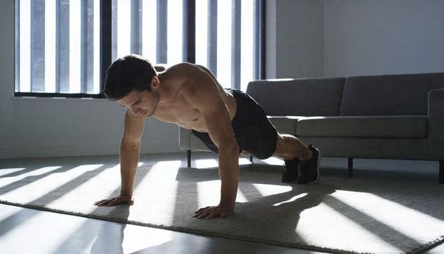 Shirtless man performing plank exercise in sunlit living room with blinds and sofa