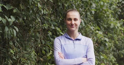 Confident woman standing arms crossed wearing lavender zip pullover against leafy wall