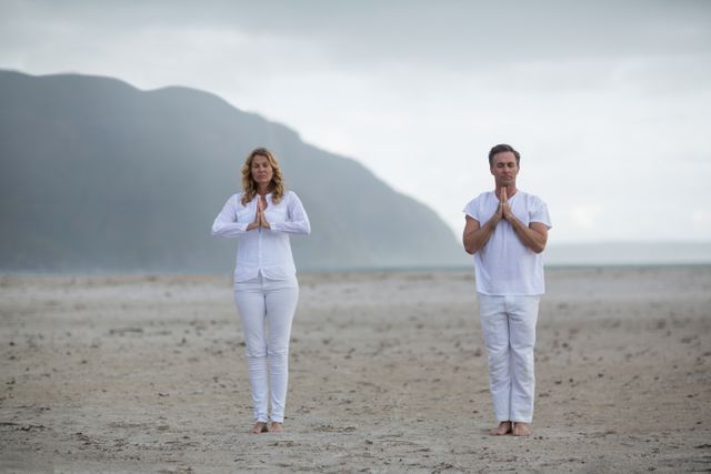 Mature couple standing on beach in white clothing, meditating with hands in prayer position. Ideal for wellness, mindfulness, and relaxation themes. Perfect for promoting yoga retreats, spiritual practices, and healthy lifestyle.