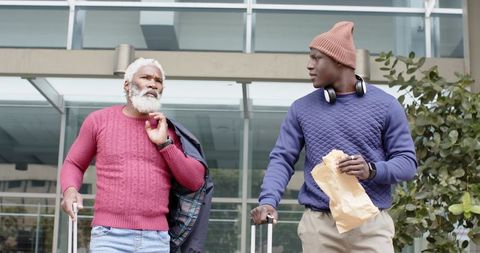 African american senior and young man walking with luggage and snack outside transit hub