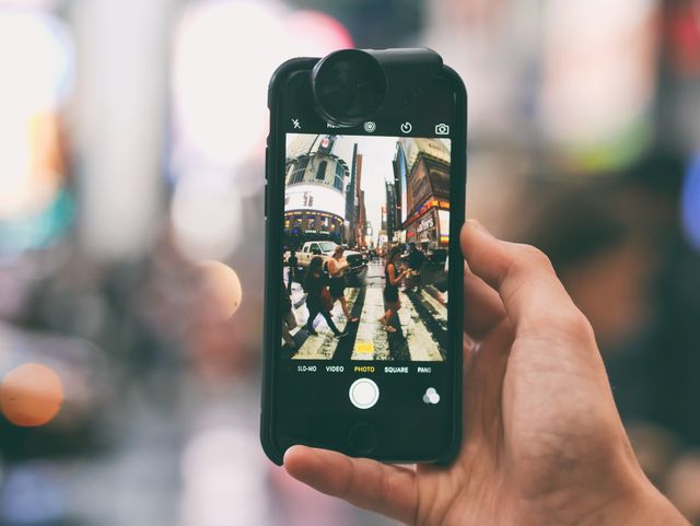 Hand holding smartphone capturing vibrant urban street scene with buildings, people, and crosswalk. Ideal for illustrating technology in daily life, urban exploration, street photography concepts, and mobile lifestyle.