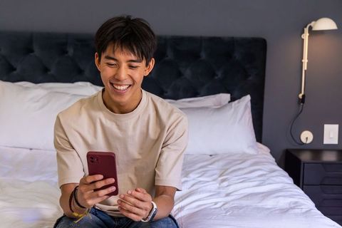Asian man smiling on bed checking smartphone and wristwatch in modern minimalist bedroom