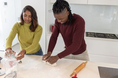 Diverse couple kneading dough and pouring flour on sunlit modern kitchen island