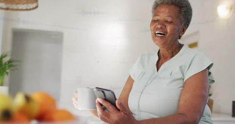 Senior woman laughing while video calling on smartphone holding mug in bright kitchen