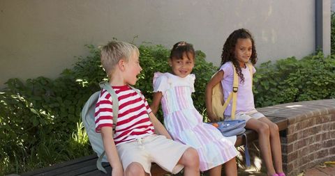 Elementary children chatting on bench with backpacks in sunny courtyard back to school