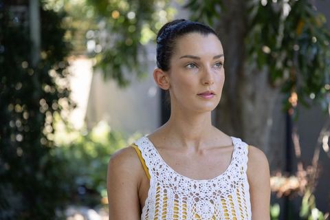 Woman standing in shaded garden wearing white crocheted top and yellow tank, bun