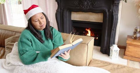 Asian Woman Reading by Fireplace in Festive Santa Hat