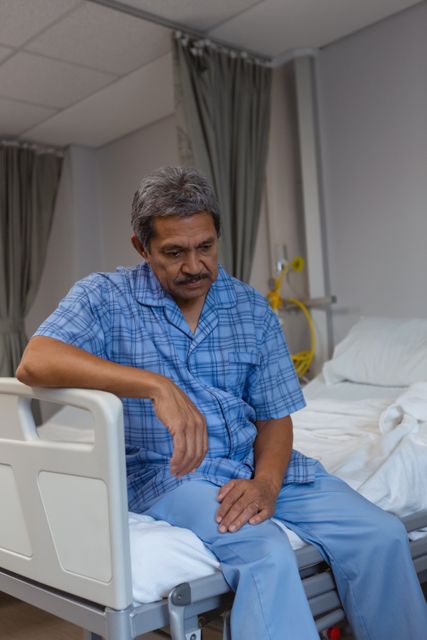 Mature male patient sitting on hospital bed, looking sad and contemplative. Ideal for use in healthcare, medical care, patient recovery, and emotional well-being contexts. Can be used in articles, blogs, and educational materials related to patient experiences, hospital care, and mental health.
