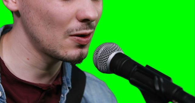 Young man singing into microphone against green screen background. Perfect for music-related content creation, video editing projects involving musical performances, visual effects, advertisements, and social media promotions. Ideal for showcasing vocal talent, audio recording, and promotional material for musicians.