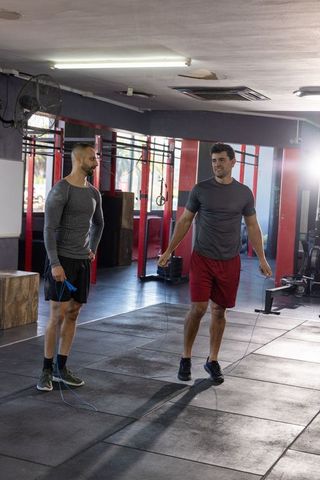 Male friends skipping rope in indoor gym training near plyo box and red rigs