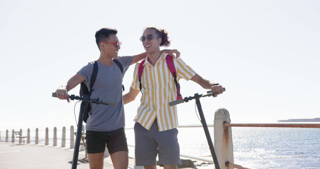 Two young men are enjoying a bright, sunny summer day at the seaside. They appear relaxed and happy as they hold their electric scooters, showcasing their camaraderie and enjoying the scenic ocean view. This image is ideal for promoting outdoor activities, travel, friendship, summer holiday destinations, and lifestyle products.