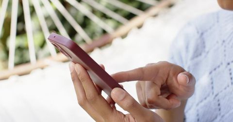 Middle-aged woman texting on smartphone while lounging in hammock wearing knit top