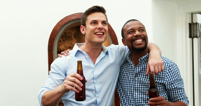 Two friends are bonding over drinks indoors. Both are holding beer bottles and smiling happily. Could be used to portray themes of friendship, celebration, joy, and leisure time. Perfect for advertisements related to social events, alcohol brands, or lifestyle and leisure concepts.
