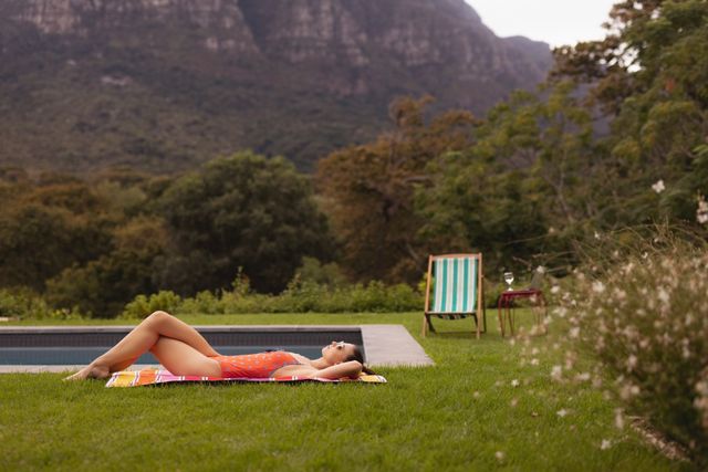 This image depicts a woman in swimwear lying on a towel near a poolside in a scenic backyard with mountains in the background. Ideal for use in travel brochures, summer vacation promotions, wellness and relaxation content, and outdoor lifestyle blogs.