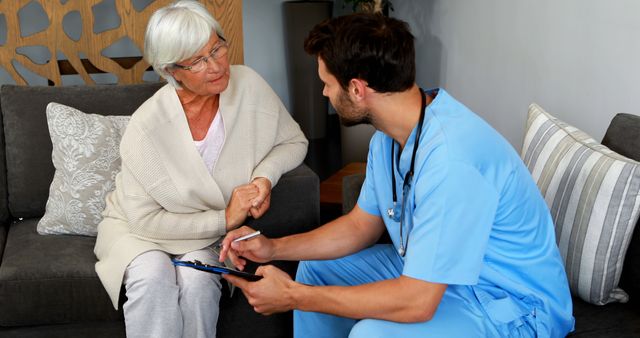 A senior Caucasian woman is engaged in a conversation with a young Caucasian male nurse, who is holding a clipboard, in a cozy home setting. Their interaction suggests a home health care visit, emphasizing the importance of medical care accessibility for the elderly.