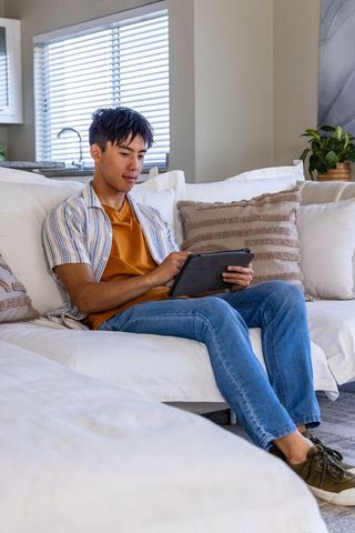 Asian man sitting on light sofa using tablet, relaxing in modern neutral living room