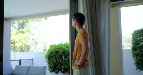 Standing asian man looking through sliding door to sunlit garden on modern patio