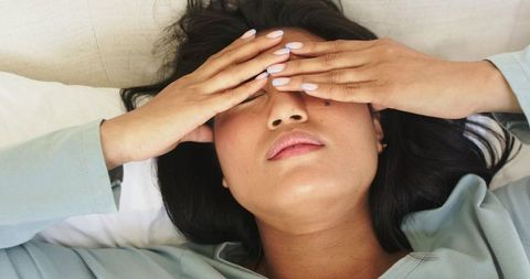 Mid-adult asian woman covering eyes while lying in bed with pastel nails and green top