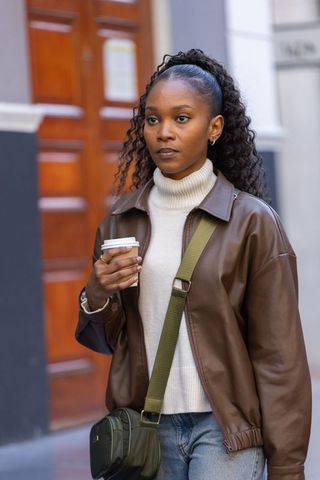 African woman walking with coffee wearing brown leather jacket and olive crossbody bag