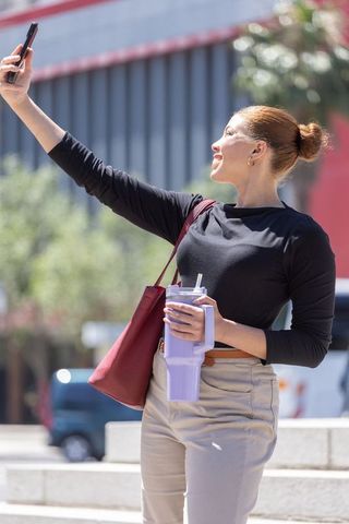 Young woman taking selfie on urban steps holding tumbler and red tote, candid lifestyle portrait
