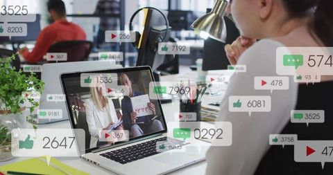 Woman watching virtual meeting on laptop with social engagement overlays in open office