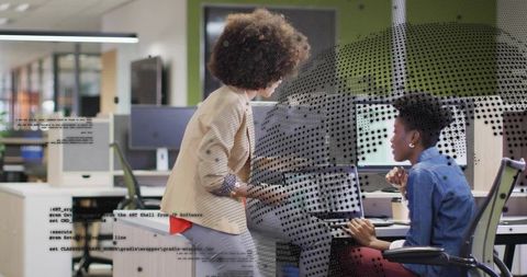 Leaning mentor guiding coworker in modern open-plan office with code overlay on monitors