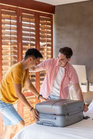 Two male friends packing gray hardshell suitcase in sunlit hotel bedroom with wooden shutters