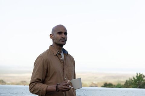 Middle-aged indian man standing on balcony holding beige mug, contemplative horizon morning