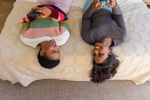 Multicultural women lying on bed holding smartphones and smiling, cozy at-home friendship
