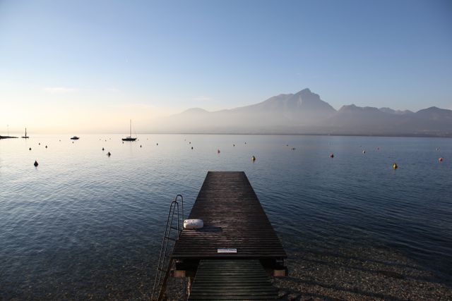 A tranquil lakeside pier extending into calm waters, surrounded by morning mist and mountains in the distance. Ideal for use in travel promotions, nature blogs, relaxation content, and inspirational posters. The scene's serenity and beauty evoke feelings of peace and contemplation.