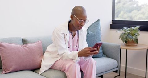 African female doctor checking smartphone sitting on sofa in modern healthcare office
