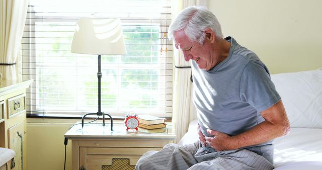 An elderly man in bedclothes sits on his bed in a bright bedroom, holding his stomach in discomfort, indicating abdominal pain or health issues. This image is suitable for illustrating concepts of health, senior care, and medical issues faced by older adults. It might be used in articles or advertisements related to healthcare services, medical conditions, and senior wellbeing.