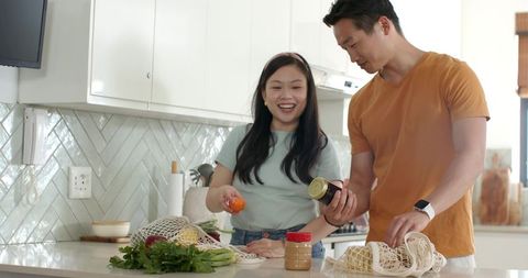 Asian couple unpacking reusable grocery bags on bright modern kitchen island