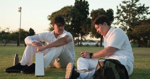 Cricketers resting on grass at golden hour discussing strategy with bat and gear