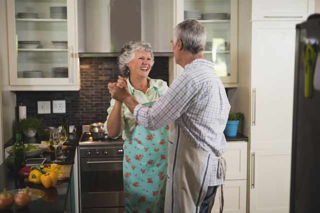 Senior couple dancing joyfully in kitchen, showcasing love and happiness in everyday life. Perfect for use in advertisements, articles, or blogs focused on senior living, healthy relationships, and home life.