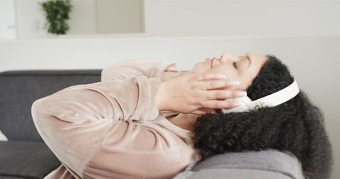 Reclining woman listening to music on white headphones on cozy gray sofa for relaxation