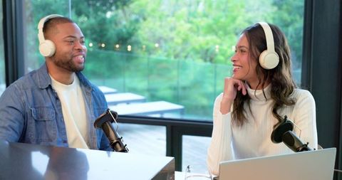 Diverse cohosts recording podcast in glass-walled studio with headphones and microphones