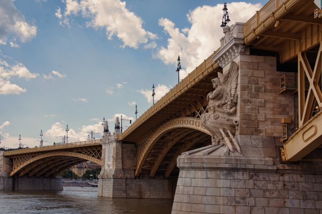 This image showcases a beautifully detailed historic bridge spanning a calm river. The bridge features intricate carvings and stonework, set against a partly cloudy sky. Ideal for promoting tourism, travel blogs, architectural studies, and urban infrastructure projects.