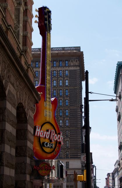 Capturing iconic guitar sign of Hard Rock Cafe in busy urban street. Sign is prominently on the side of a historic brick building, with skyscrapers visible in background. Suitable for travel advertisements, city guides, tourism brochures, promoting nightlife spots and popular restaurants.