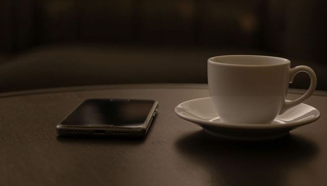 White ceramic cup on saucer with smartphone on dark wood table, cozy cafe mood, warm lighting